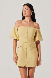 A woman is wearing a light yellow, off-the-shoulder dress with large, puffed short sleeves. The dress has a row of buttons down the front and two flap pockets at the waist.